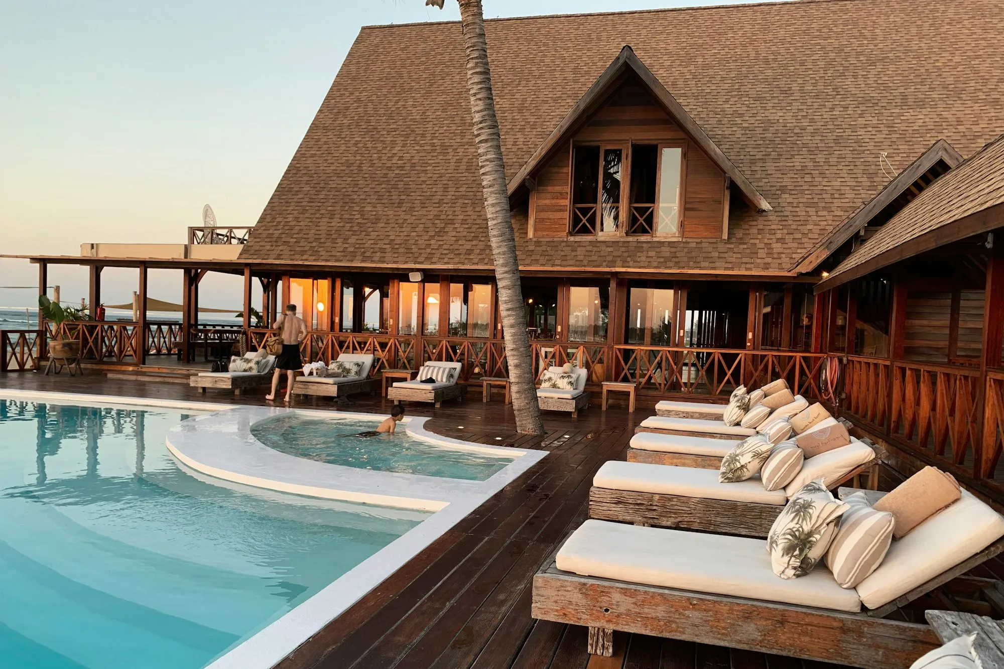 Loungers by a pool in front of a wooden beach house at sunset